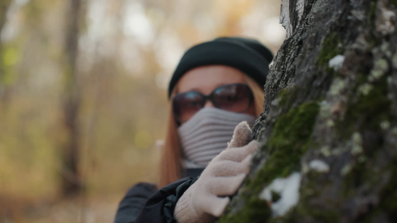 Donna bianca mascherata sbircia da un albero muschioso, mano guantata sulla corteccia. Sfondo di foresta autunnale con profondità di campo ridotta. Movimento furtivo che suggerisce una ladra o un'intrusa che perlustra l'area, in luce soffusa e un clima freddo.