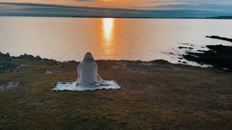 Aerial-of-beautiful-young-woman-traveler-sitting-on-blanket-in-front-of-Atlantic-Ocean-in-France,-Brittany-during-sunset-golden-hour