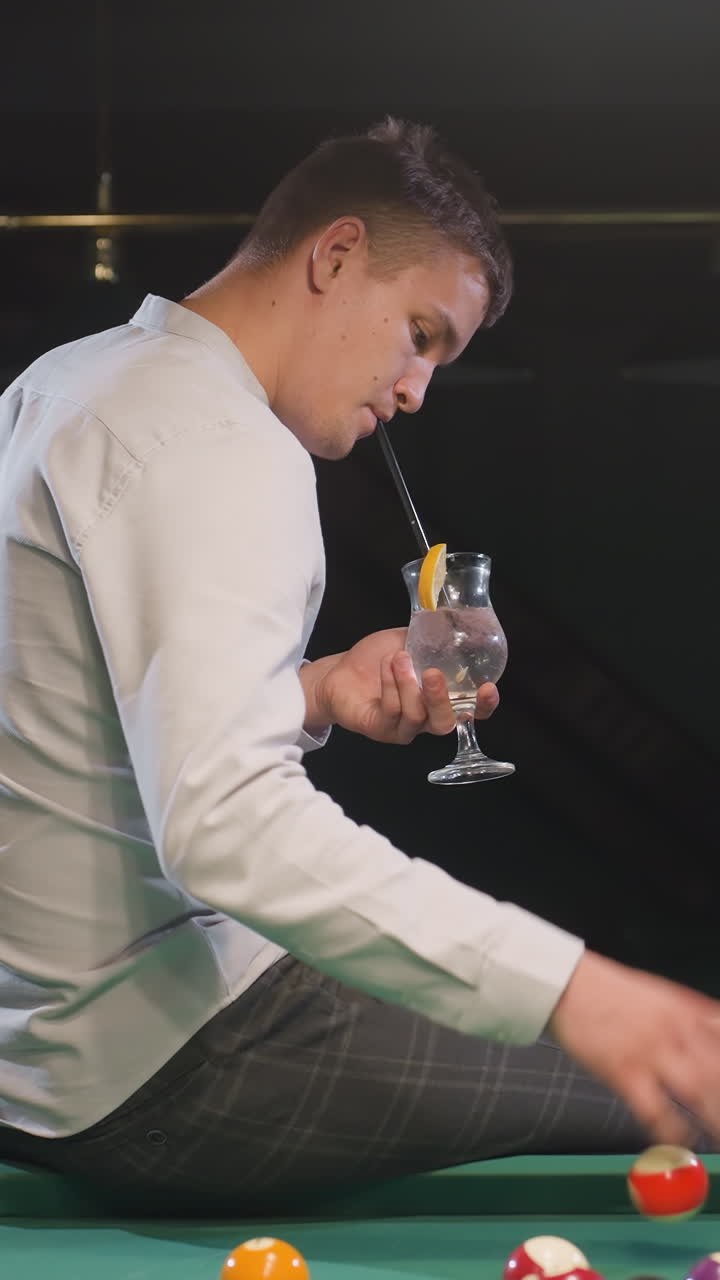 색 셔츠와 체크형 바지를 입은 신사님이 초록색 빌리어드 테이블에 앉아서 리몬 음료 한 잔을 들고 빌리어드를 고 있습니다. 비언트 조명과 현대적인 빌리어트 홀과 함께 세련된 설정