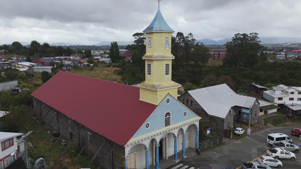Aerial view capturing the vibrant Iglesia de Nuestra Senora del Rosario, surrounded by Chonchi's traditional wooden houses and lush greenery