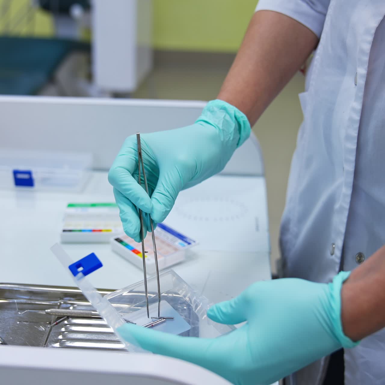 Person in white coat and blue latex gloves holds metal forceps. Metal nozzles are taken from a plastic box and put into the steel tray on the drawer. Close up