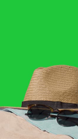 Vertical-Video-Summer-Holiday-Concept-Of-Sunglasses-Sun-Hat-Camera-Beach-Towel-On-Sand-Against-Green-Screen