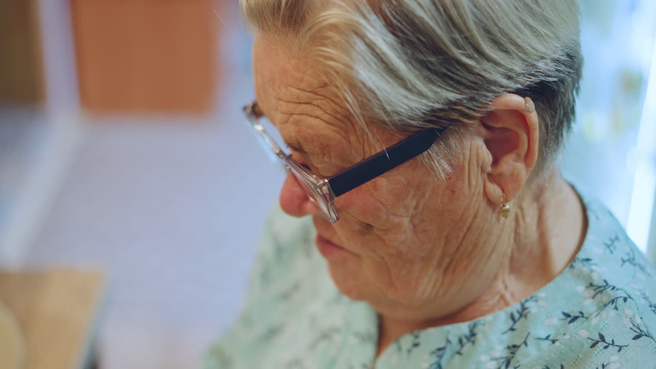 Primo piano di una nonna che esamina una ricetta al tavolo della cucina. Indossa un vestito floreale e gli occhiali da lettura, ha un'espressione concentrata. Le mani sono vicino a una ciotola e un cucchiaio, nella calda luce del mattino, in un momento nostalgico di vita casalinga.