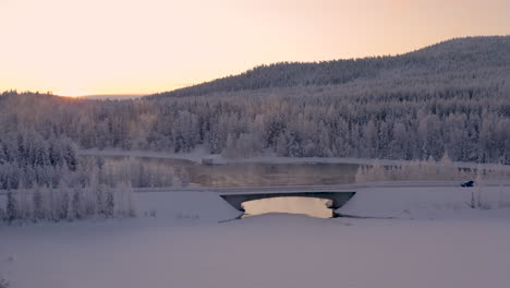 aerial view flying across wintertime woodland with vehicle driving along freezing misty ice lake bridge