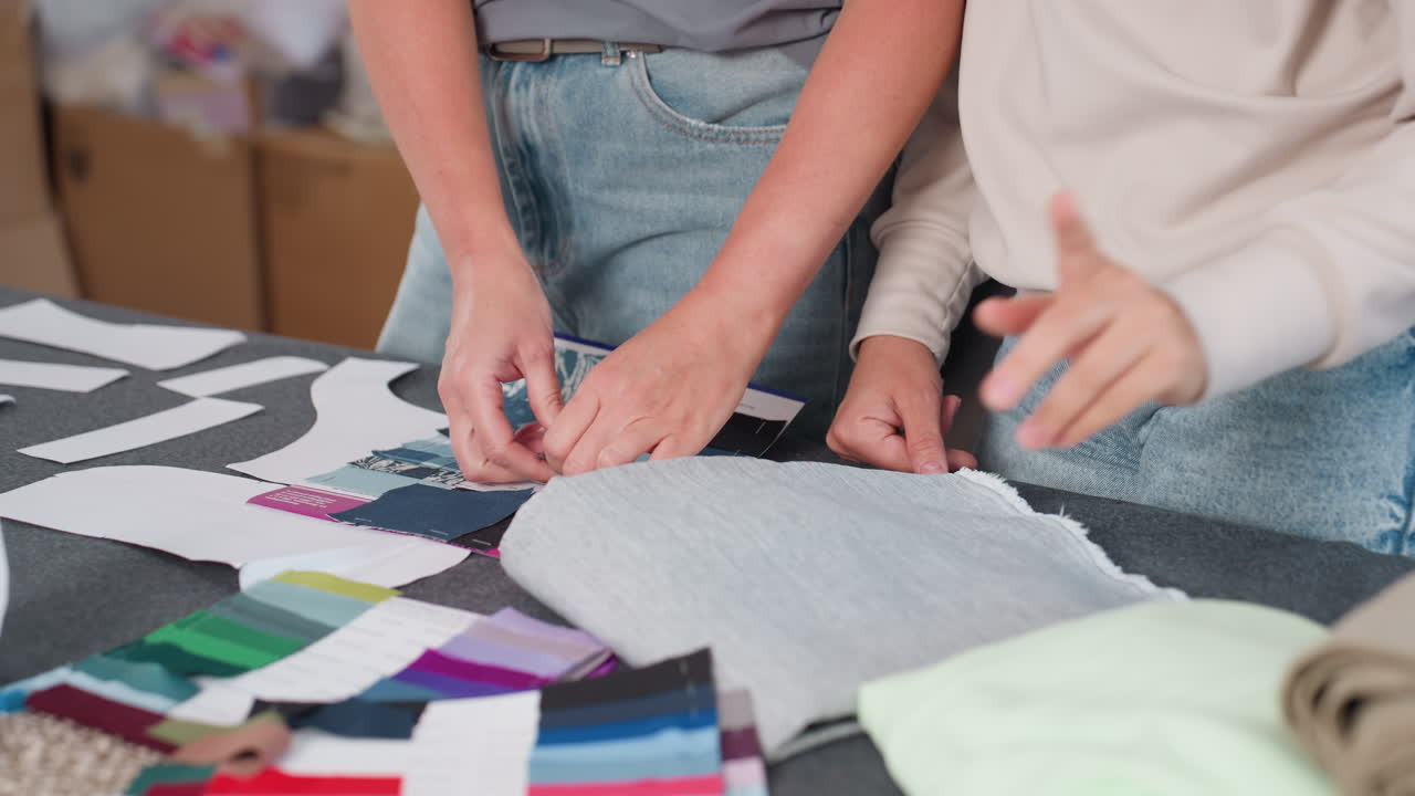 Beautiful women in jeans selecting colorful fabrics on creative workspace table, one pulling out plain material by corner while reviewing textile samples, surrounded by folded cloths