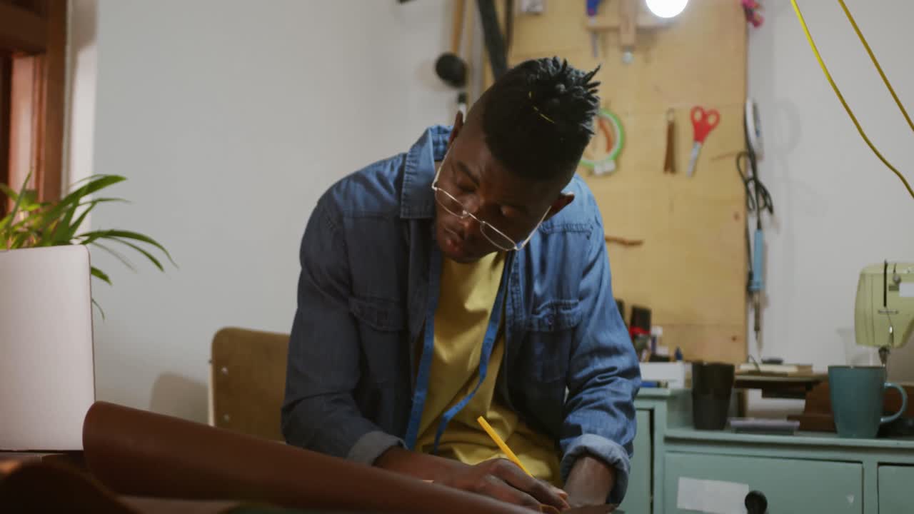 Thoughtful african american craftsman wearing glasses making notes in leather workshop