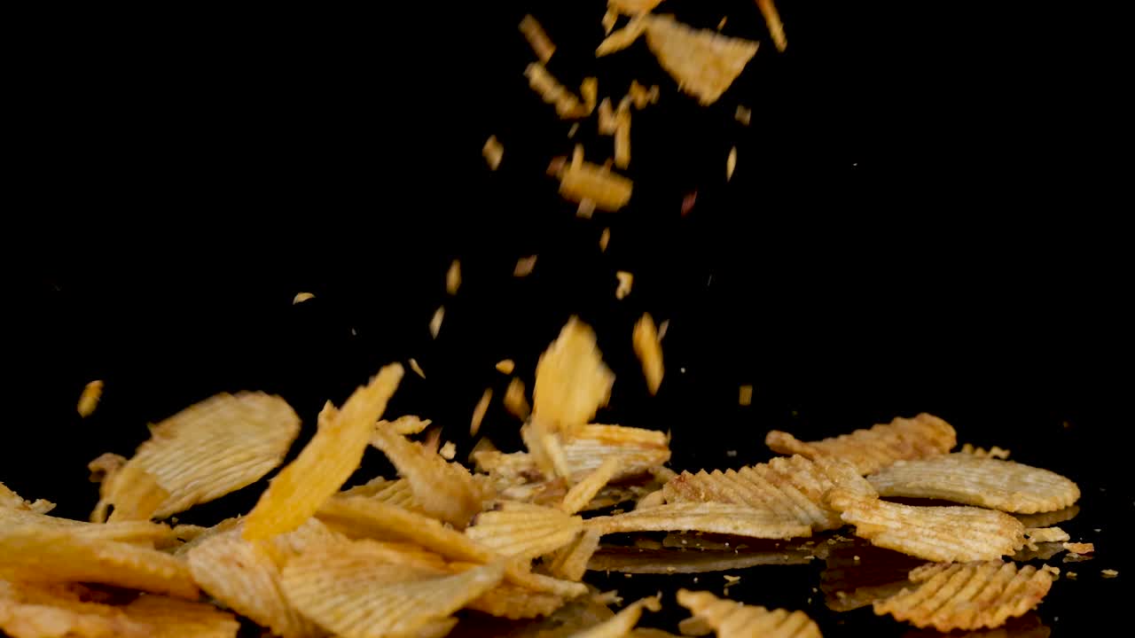 Wavy potato chips fall and scatter onto a glossy black surface, captured in high-speed slow motion with dramatic lighting and a dark background