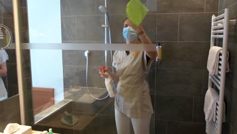 a housekeeping employee with face mask is cleaning and disinfecting the shower of a hotel room