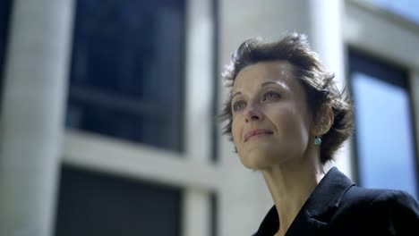 close-up of thoughtful businesswoman standing outdoors in street and looking away while waiting for somebody, her short hair waving in wind