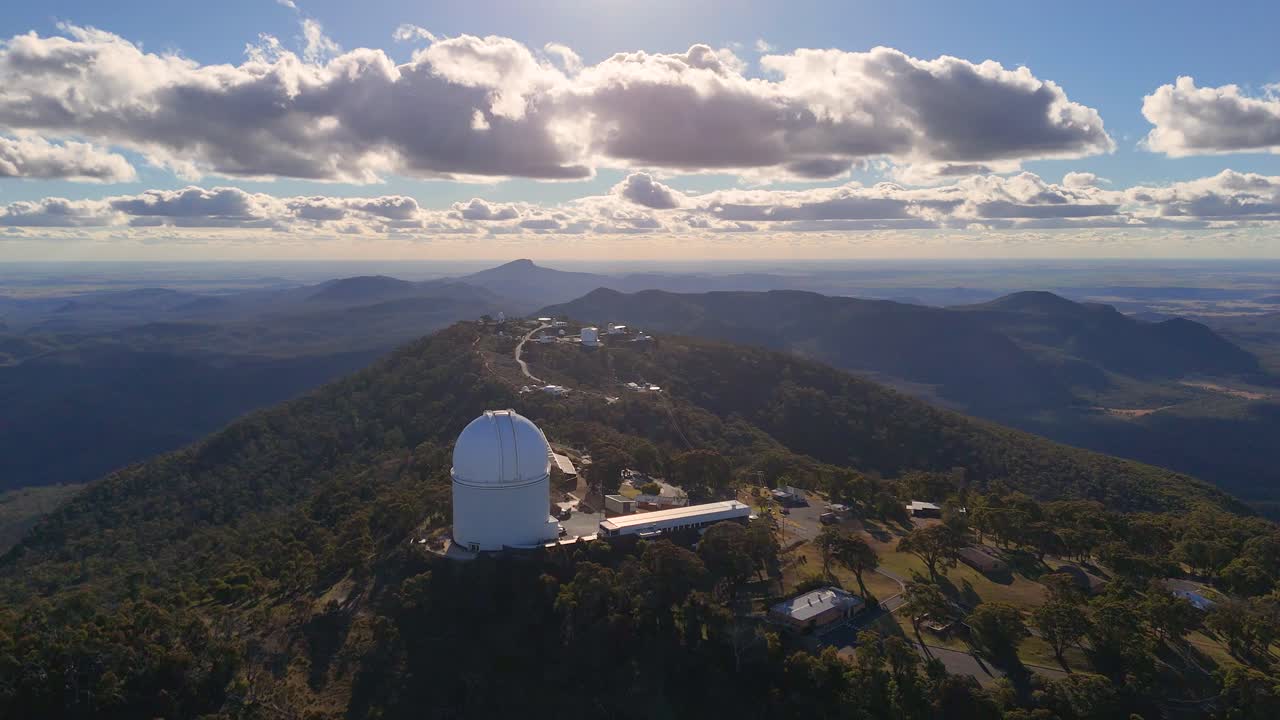Aerial footage captures a large observatory dome atop a forested mountain ridge, with dramatic clouds and golden hour sunlight, filmed in Coonabarabran, NSW