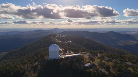 aerial footage captures a large observatory dome atop a forested mountain ridge, with dramatic clouds and golden hour sunlight, filmed in coonabarabran, nsw