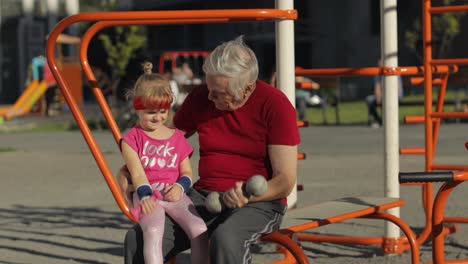 Granddaughter-and-grandfather-doing-fitness-exercises-with-dumbbells.-Senior-man-with-child-kid-girl