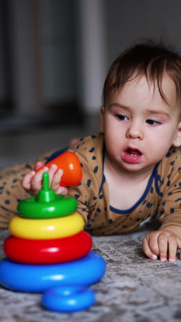 baby boy in brown romper lies on the carpet indoors. adorable one-year old child playing with a toy pyramid. blurred backdrop. vertical video