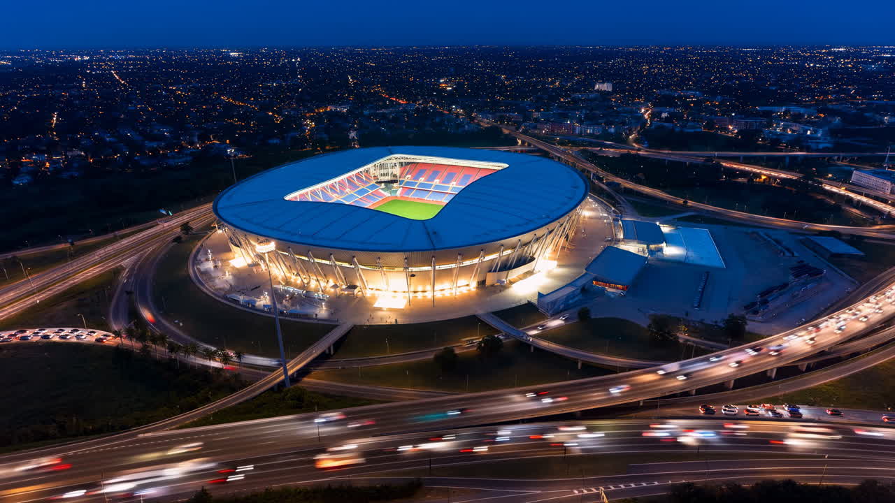 Nocny stadion z lotu ptaka, oświetlona arena, nowoczesny okrągły stadion z podświetlanym dachem i żywą murawą, otoczenie.