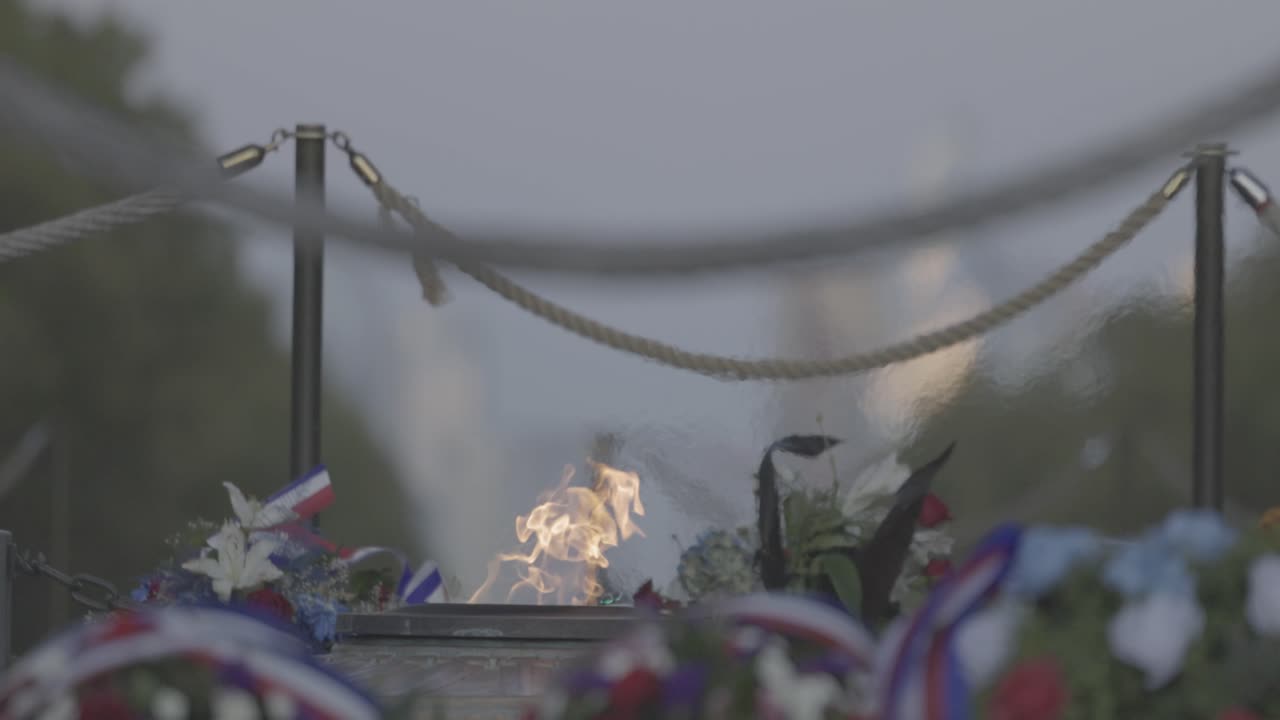 Primo piano di una fiamma eterna che tremola presso un sito commemorativo all'aperto, circondato da gigli bianchi e fiori commemorativi. Una barriera di corda incornicia la scena solenne sotto un cielo alpino coperto.