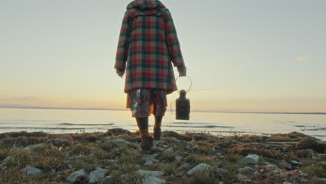 woman with lantern walking on shore towards lake at sunset