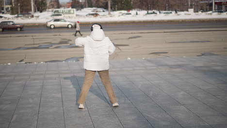 back view of elegant woman wearing winter coat and headphones dancing alone on wide tiled walkway near busy two way road with passing vehicles and pedestrians in snowy urban landscape