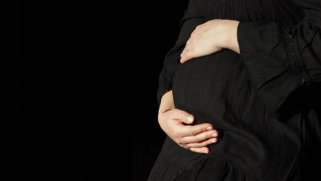 a pregnant woman gently holds and rubs her round belly with copyspace against a black background