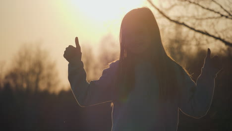 Ein-Junges,-Glückliches-Mädchen,-Das-Zwei-Daumen-Nach-Oben-Zeigt,-Während-Es-Sich-Vor-Dem-Sonnenuntergang-Abhebt