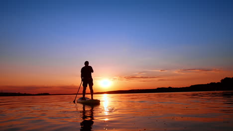man practicing sup boarding on the river at sunset. low angle view on the person standing on the board and rowing with an oar. low angle view.
