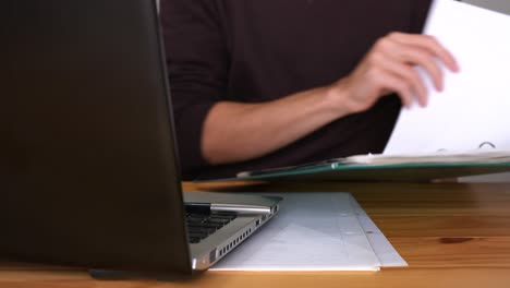 oficinista masculino mirando archivos en una carpeta verde con laptop y papeles en una mesa de oficina de madera