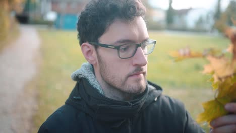 Slow-motion-capture-of-young-European-male-examining-autumn-leaves-on-trees,-realizing-winter's-chill-is-near