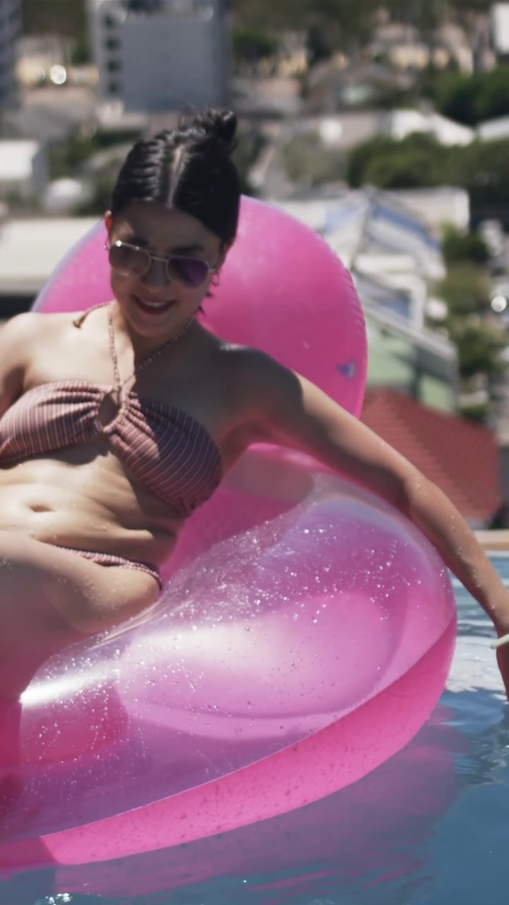 Vertical video: Leaning woman in bikini on pink float in roof pool, smiling toward man appearing