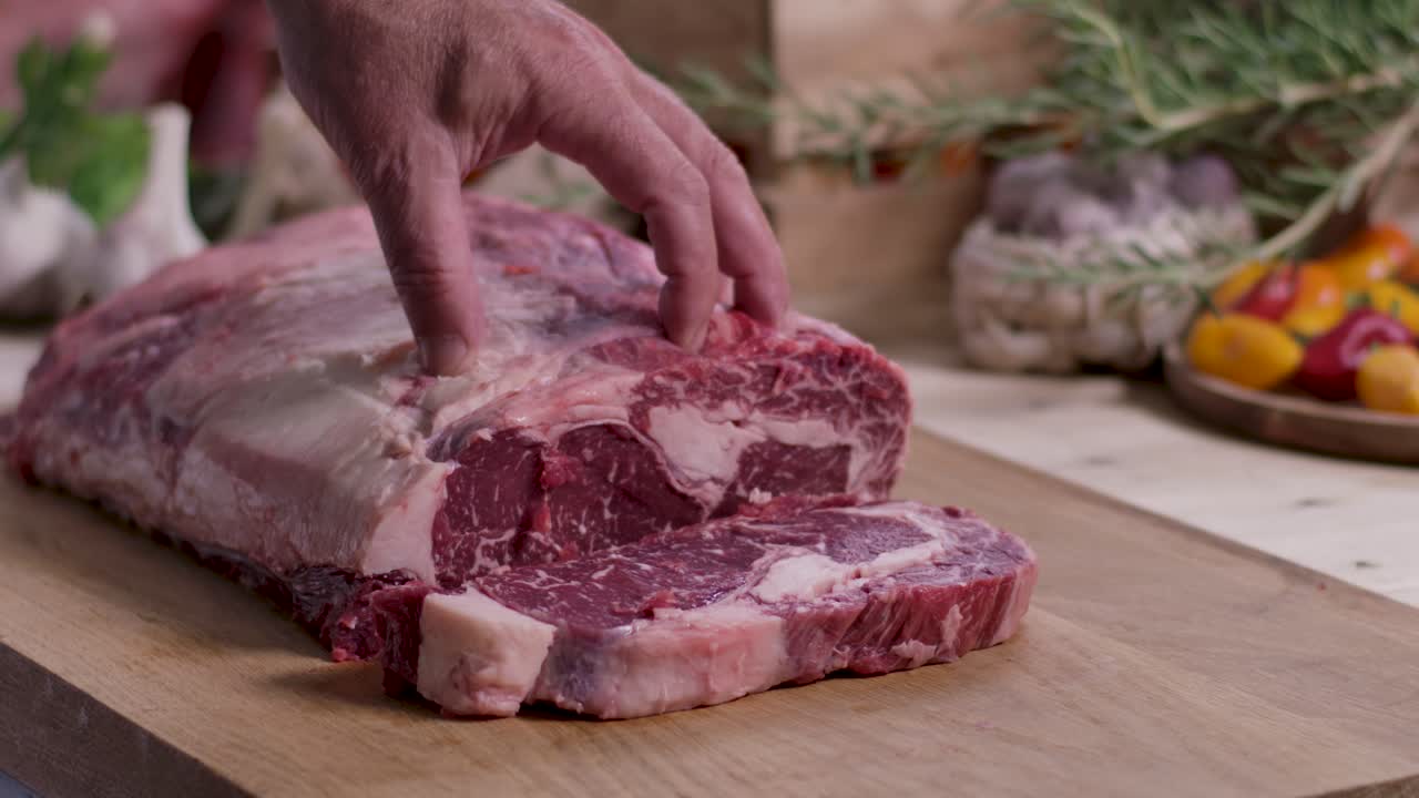 Close up side view of a professional chef using a sharp knife to carve a marbled ribeye steak from a large raw beef roast on a wooden board