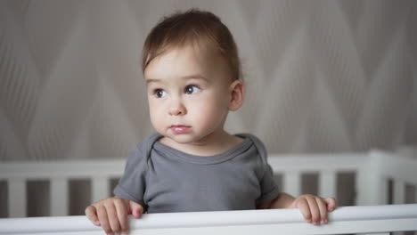 sweet adorable baby standing in his crib after waking up. calm caucasian toddler in the bedroom. white blurred backdrop.