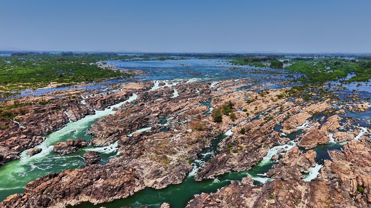 Expansive aerial view of the Mekong River at Don Det, Laos, where winding rapids carve through rugged rocky terrain and patches of dense green vegetation spread across the wide flowing water
