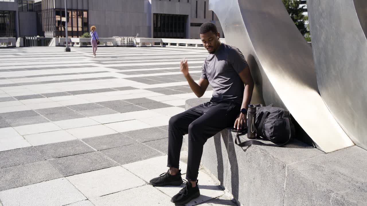 African American man glancing left in plaza touching earbud taking phone checking texts pack