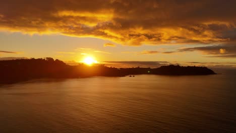 stunning sunset scene at onetangi beach on waiheke island, new zealand, captured during the golden hour. the sky is painted with dramatic clouds as the soft orange light reflects over calm sea.