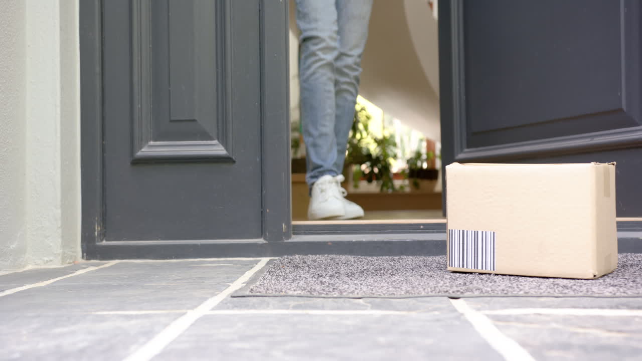 Parte inferiore di un uomo birazziale che raccoglie una scatola di cartone fuori dalla porta d'ingresso, spazio per il testo, al rallentatore.