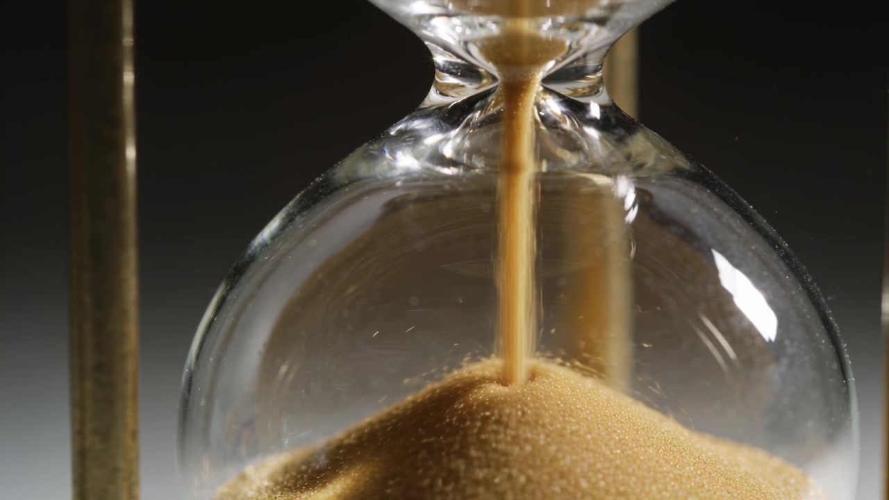 Steady thin stream feeding hourglass lower bulb, golden sand forming conical mound, studio lighting