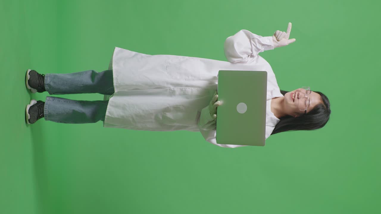 cuerpo lleno de mujer científica asiática con una computadora portátil sonriendo y señalando a un lado mientras está de pie en el fondo de la pantalla verde en el laboratorio