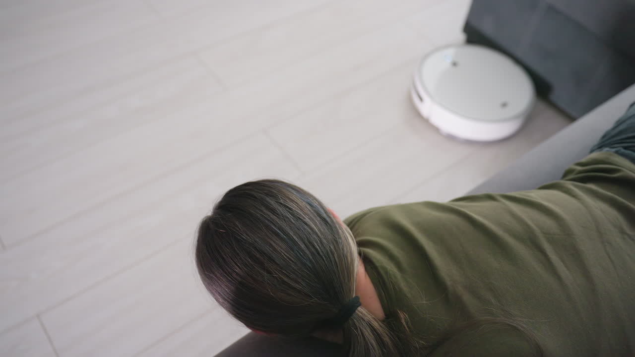 Top down view of woman with long tied hair lying on sofa watching robot vacuum move across floor, representing relaxation, leisure, home cleaning technology