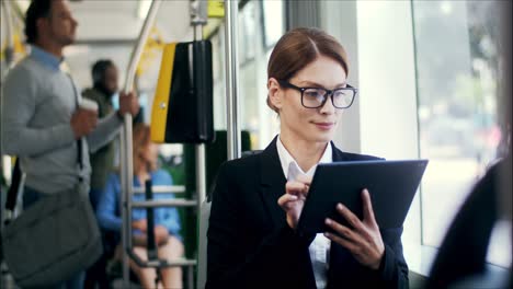 Good-Looking-Young-Woman-In-Official-Style-And-Glasses-Sitting-In-The-Tram-While-Going-Home-And-Tapping-Or-Scrolling-On-The-Tablet-Computer