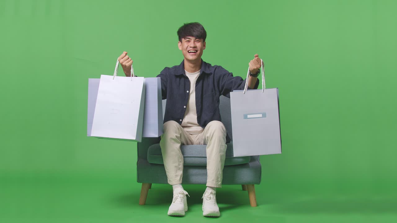 녹색 스크린 앞에 소파에 앉아 쇼핑 가방을 들고 있는 아시아 쇼핑맨의 앞면