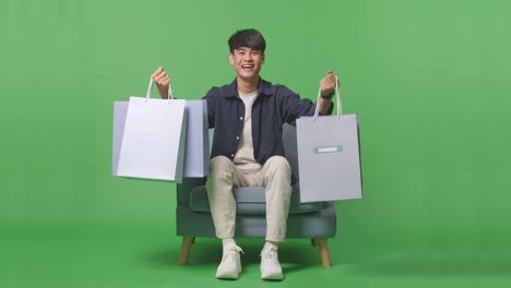 vista frontal de un hombre de compras asiático sosteniendo bolsas de compras sonriendo a la cámara mientras está sentado en el sofá frente a la pantalla verde