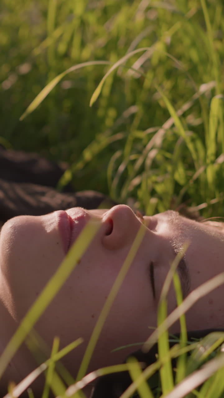 primer plano de una mujer joven acostada en un campo de hierba iluminado por el sol, perdida en sus pensamientos, rodeada de alta hierba verde que enmarca suavemente su rostro, el cálido resplandor de la luz del sol en el fondo