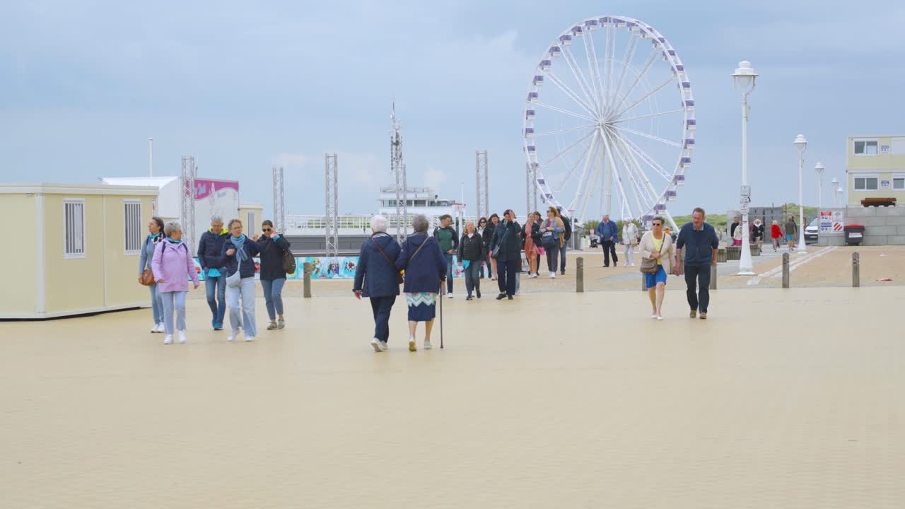 People enjoying relaxed day on sea promenade near the beach of Nieuwpoort with ferris wheel in background. Holiday town by the Belgian sea - Slow motion
