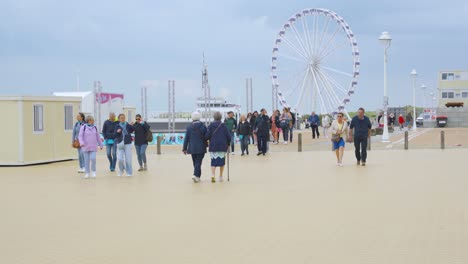 people enjoying relaxed day on sea promenade near the beach of nieuwpoort with ferris wheel in background. holiday town by the belgian sea - slow motion
