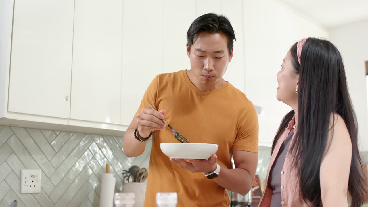 白いボウルとスプーンで味付けを確認するために、料理を味見する男性と、キッチンカウンターにいるアジア人カップル。