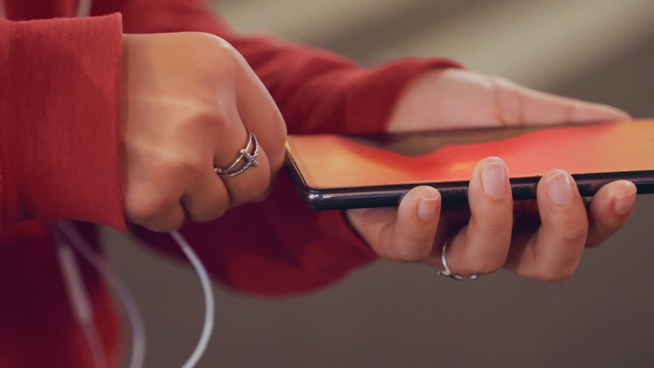 Primo piano di mani che regolano la playlist dello smartphone. Indossa una felpa rossa con cappuccio, auricolari inseriti, un delicato scorrimento sullo schermo. Incrocio sfocato e luci soffuse sullo sfondo, anelli e postura rilassata, una serata intima.