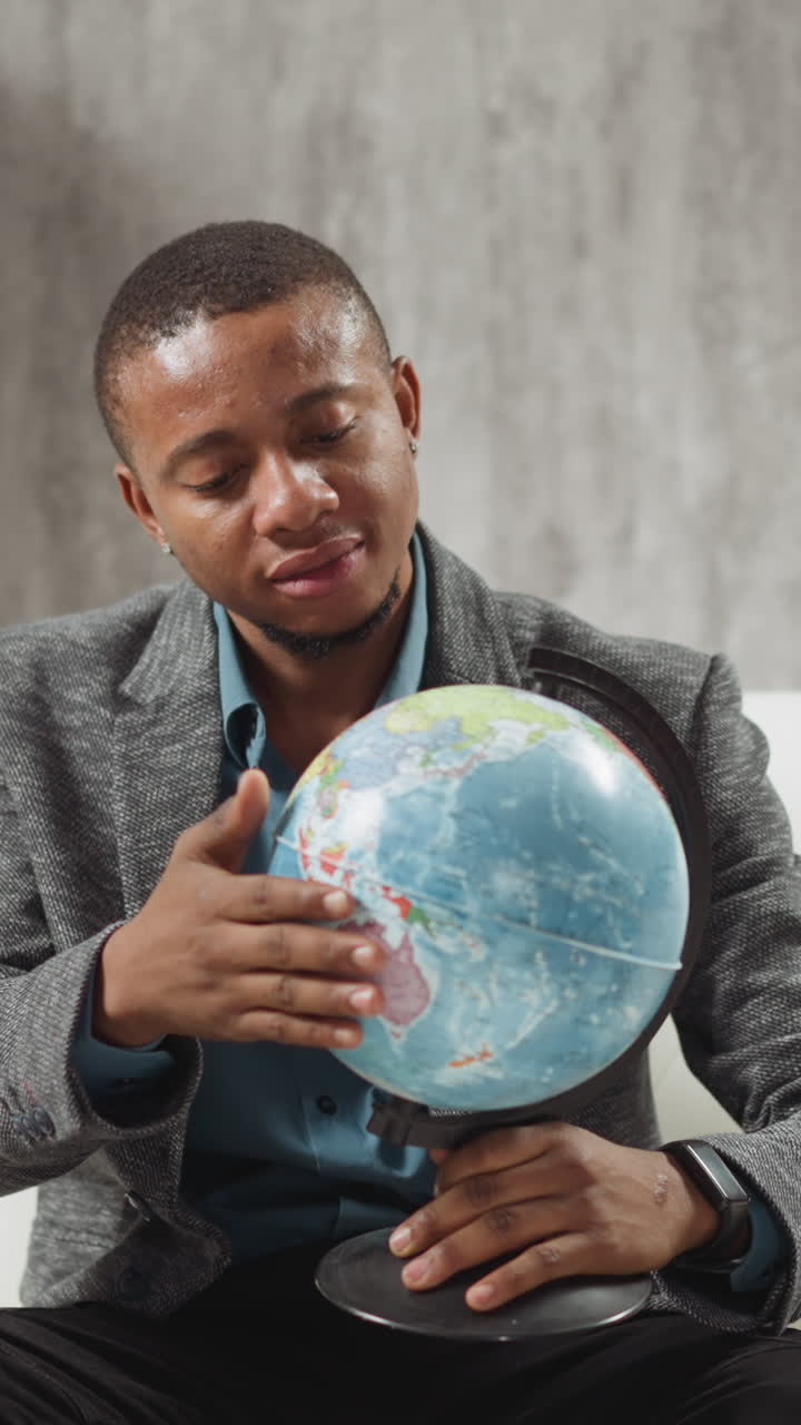 Cheerful young African-American man geography teacher turns globe sitting on couch near open laptop ready for online lesson at home slow motion