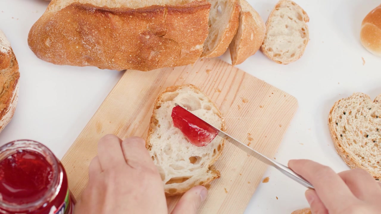 High angle shot of hands spreading red fruit jam onto crusty bread on wooden board.