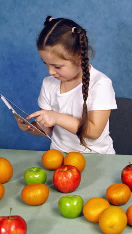 little girl holding tablet in the kitchen. sweet fruits laying on the table. cute child playing games on gadget near the table with fresh apples and oranges. vertical video