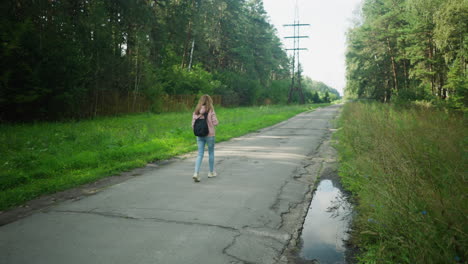 back view of student walking alone with head bowed on cracked tarred road flanked by green forest, distant power cables, and roadside puddles under daylight, conveying solitude and introspection