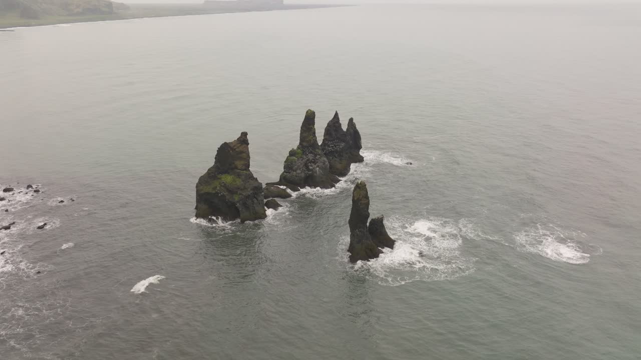 Aerial view of the iconic Reynisdrangar sea stacks off the coast of Reynisfjara, Iceland, rising dramatically from the ocean amidst crashing waves.