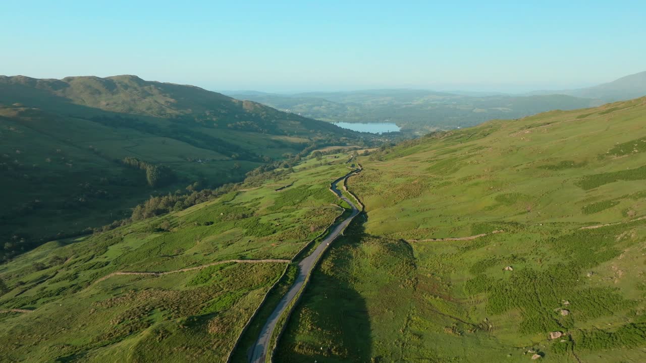 Mountainside winding road illuminated by early morning sun with Lake Windermere in the distance. Kirkstone Pass, Lake District, Cumbria, UK